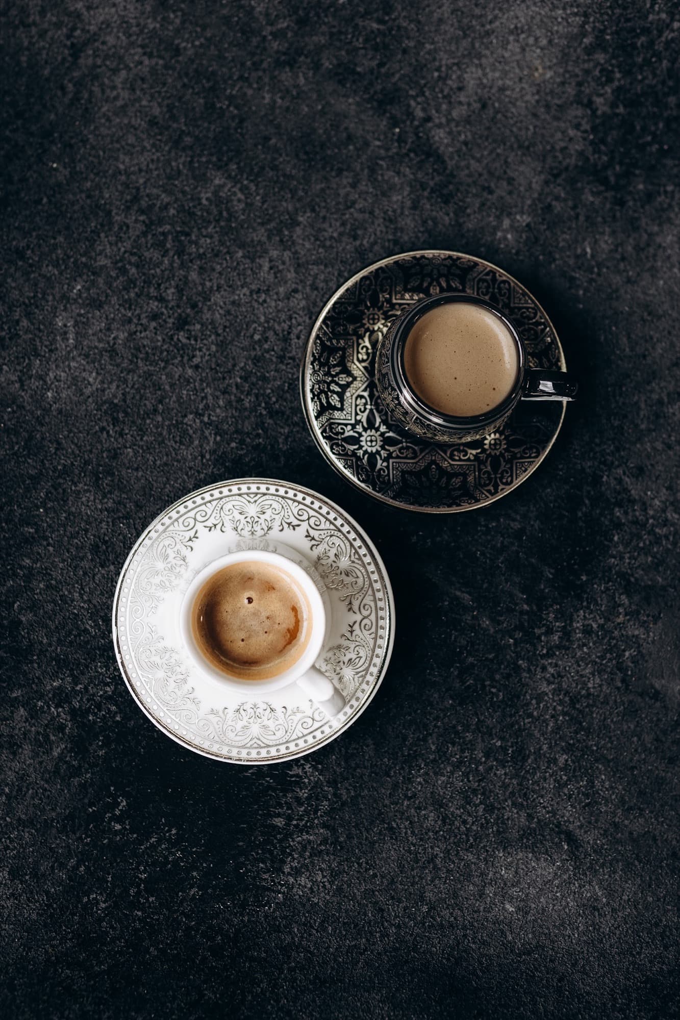Two cups of Arabiya coffee shot from above on a dark stone surface — one in an ornate silver-and-black saucer and one in a white porcelain cup, espresso crema gleaming.