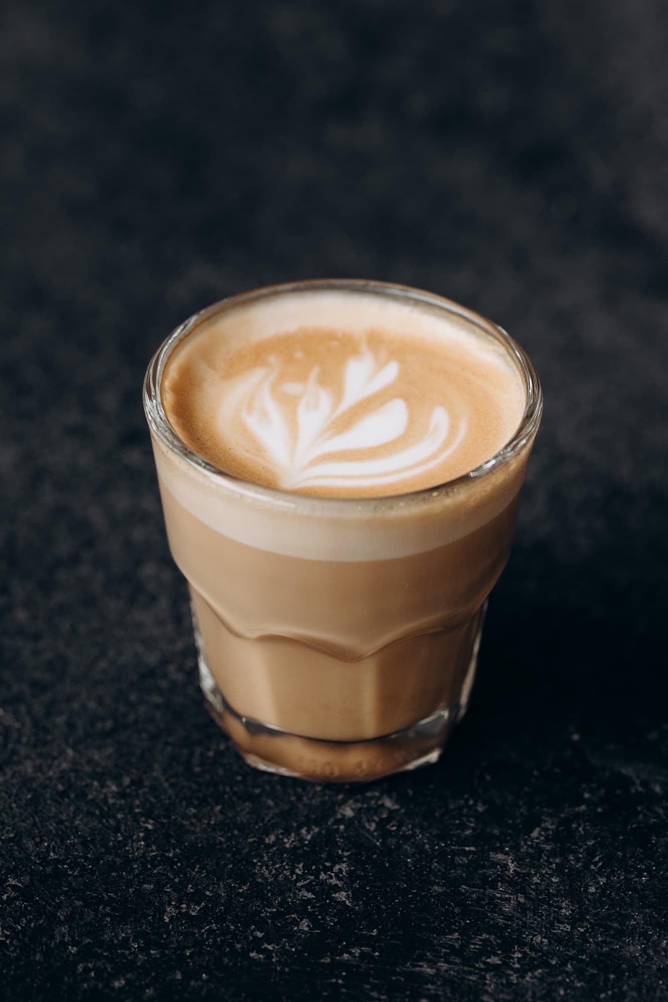A cortado in a small clear glass on a dark stone surface — espresso met by steamed milk, finished with a heart-and-leaf latte-art rosetta.