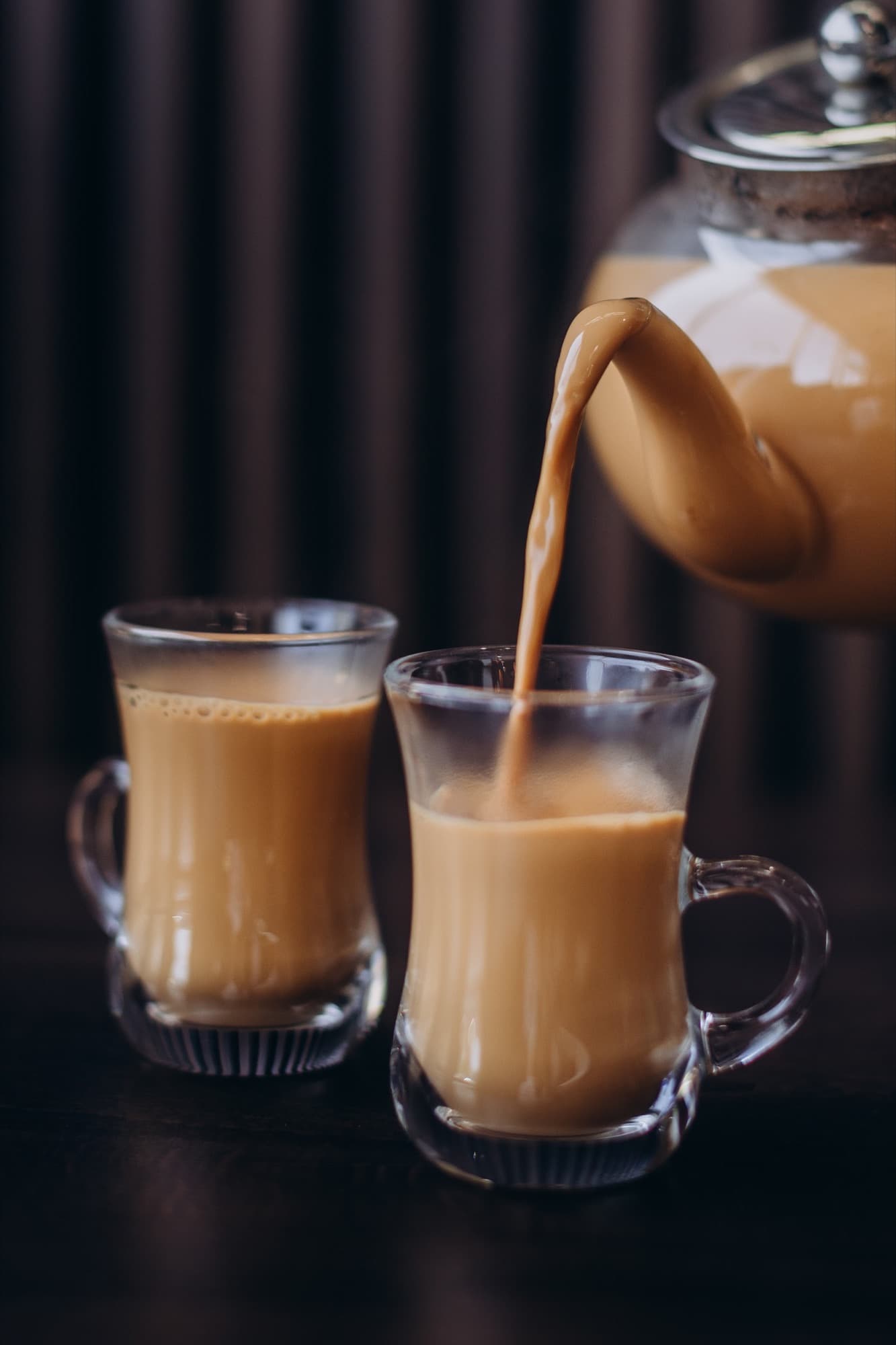 Karak tea being poured from a teapot into a clear glass cup — warm caramel-amber color, the second cup waiting alongside.