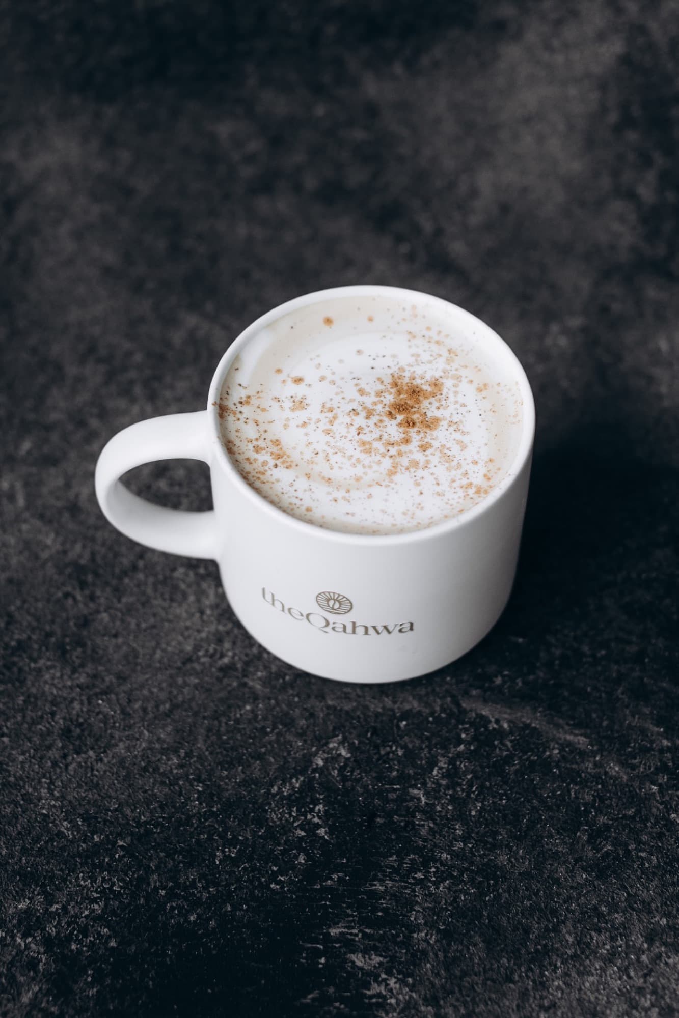 Hot Spanish Latte in a branded theQahwa ceramic mug — silky microfoam dusted with cinnamon on a moody dark stone surface.