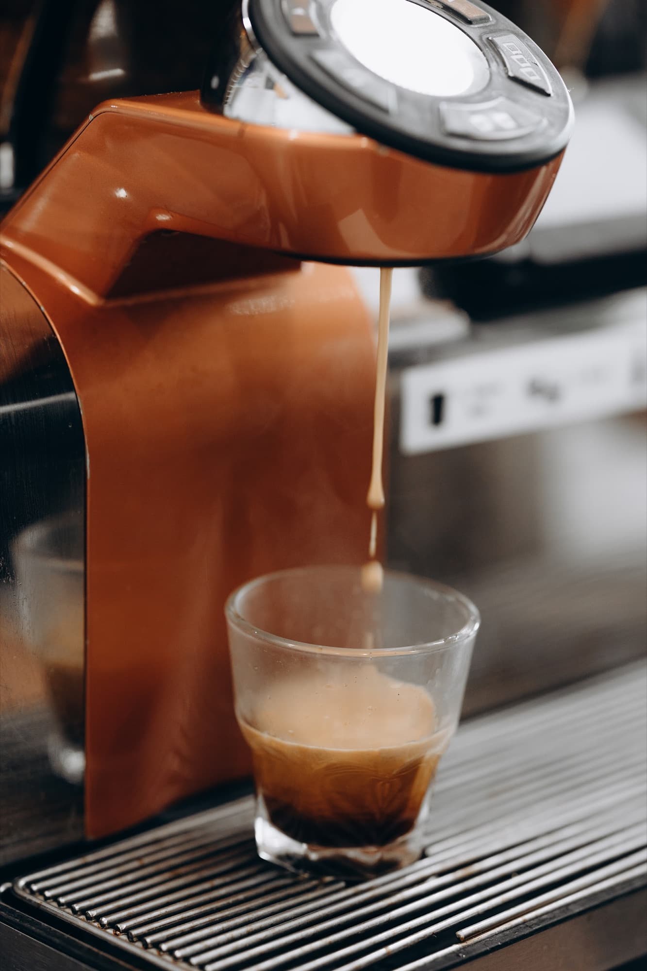 Espresso pulling into a small clear glass on the drip tray of a copper-orange La Marzocco — golden crema streaming down from the group head.