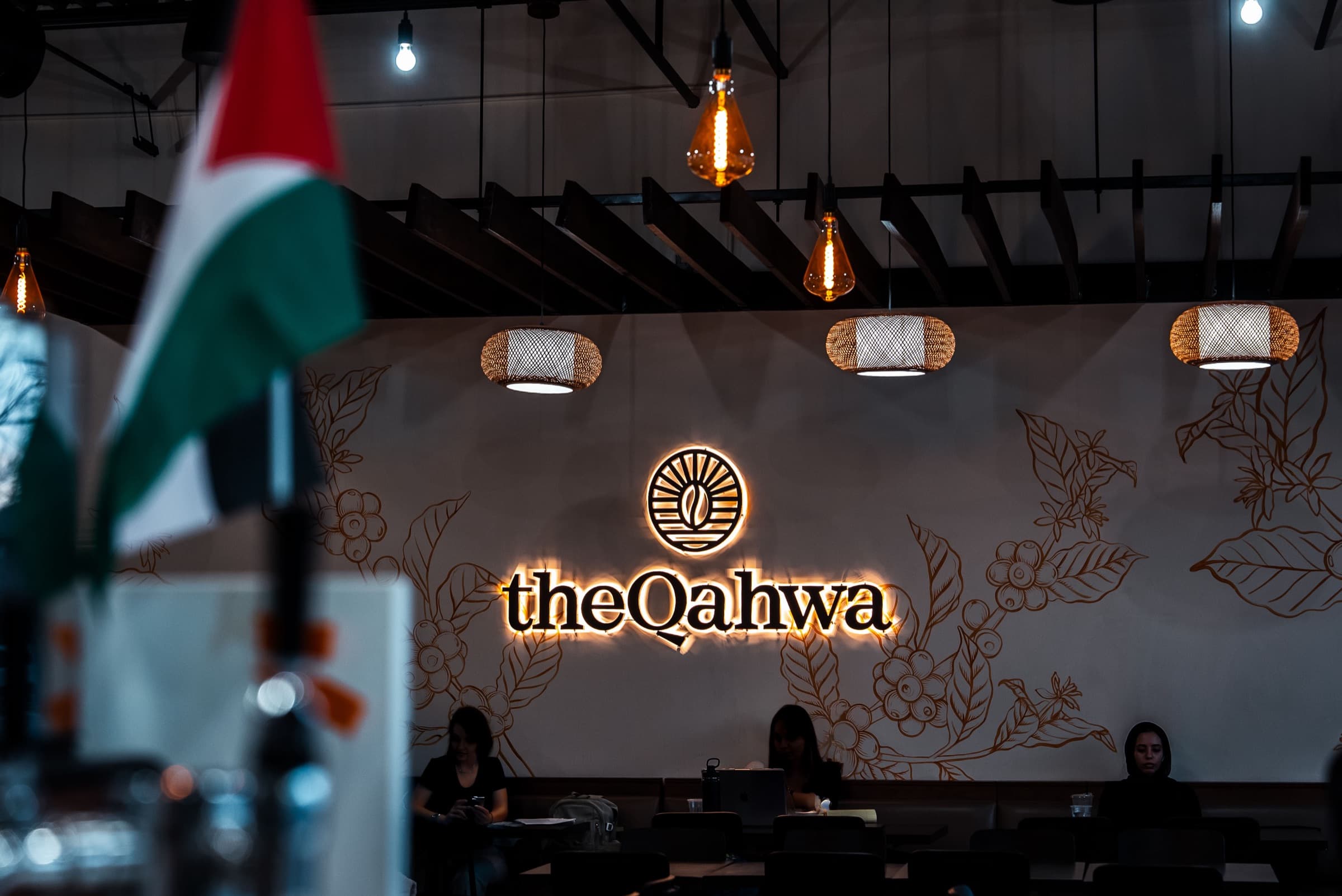 The Qahwa Bridgeview café — the illuminated theQahwa wordmark glowing against a coffee-plant mural, with warm pendant lights overhead.