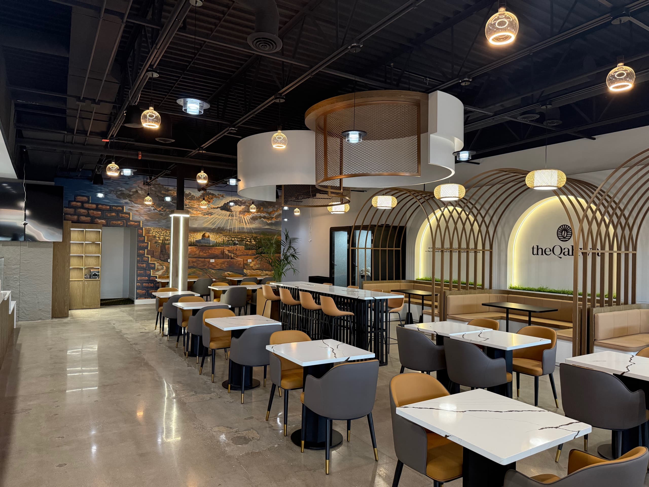 The Qahwa Orland Park interior — arched bronze trim with the brand mark, custom pendant lighting, and a hand-painted mural across the back wall.